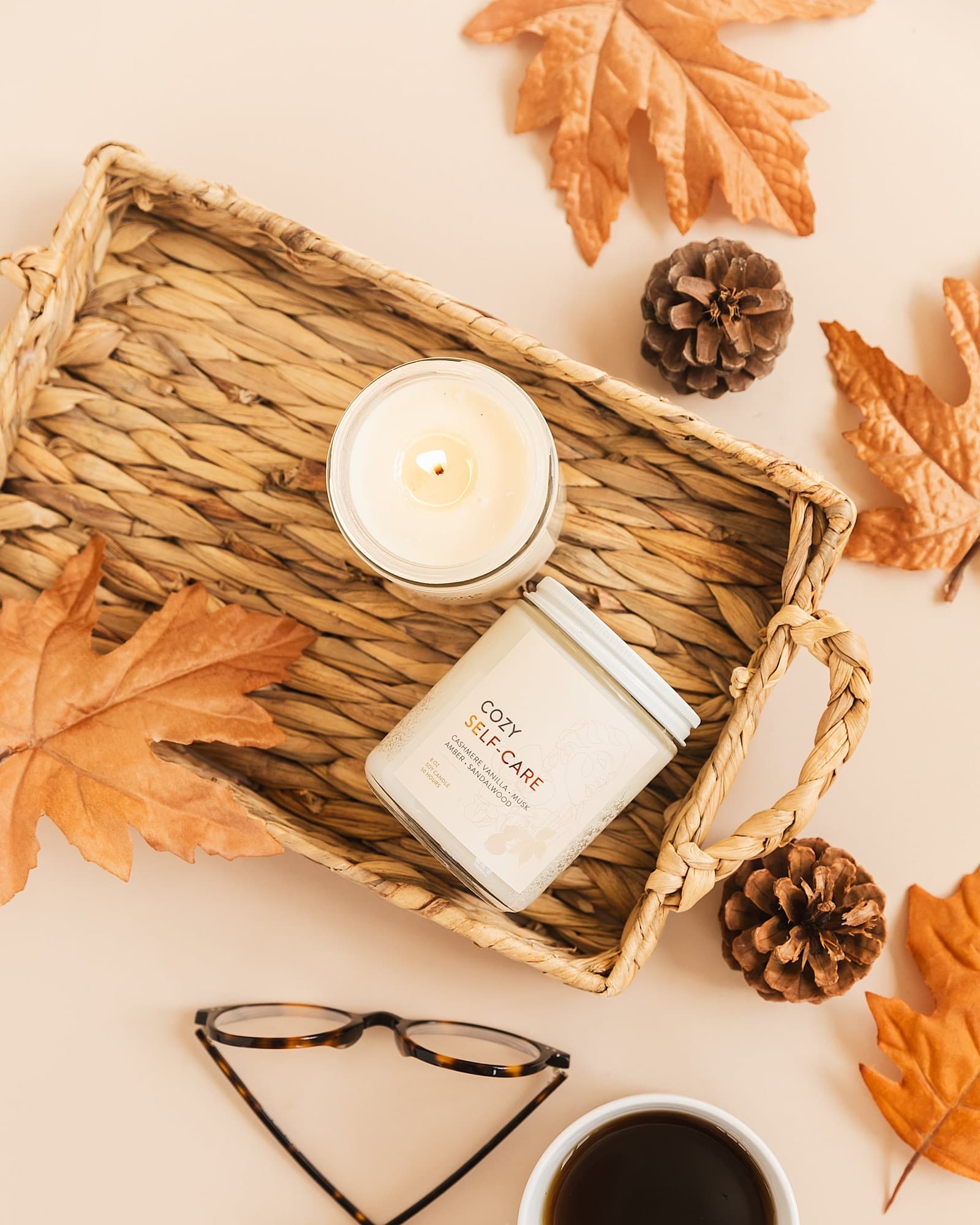 A lit Ignite Your Peace candle on a woven tray with autumn leaves, glasses and coffee